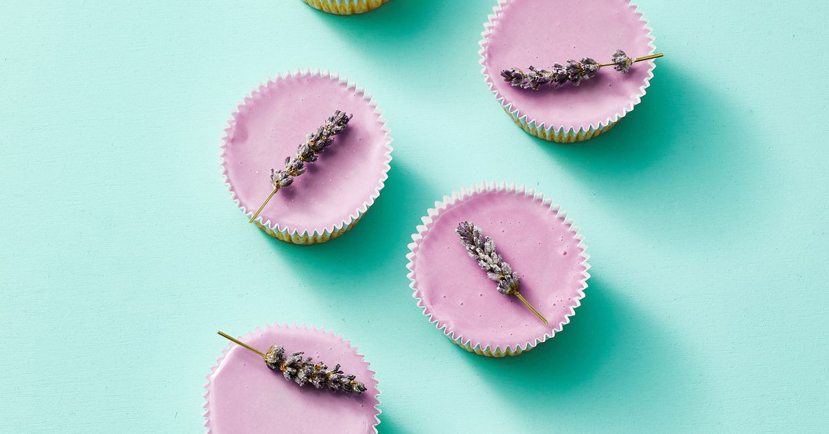 four lavender colored cupcakes topped with candied pieces of lavendar on a light blue backdrop