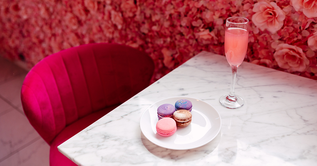 rose-filled wall with a pink velvet chair sitting near a marble table top. there is a white plate with four macarons on it and a champagne flute filled with a pink mimosa. 