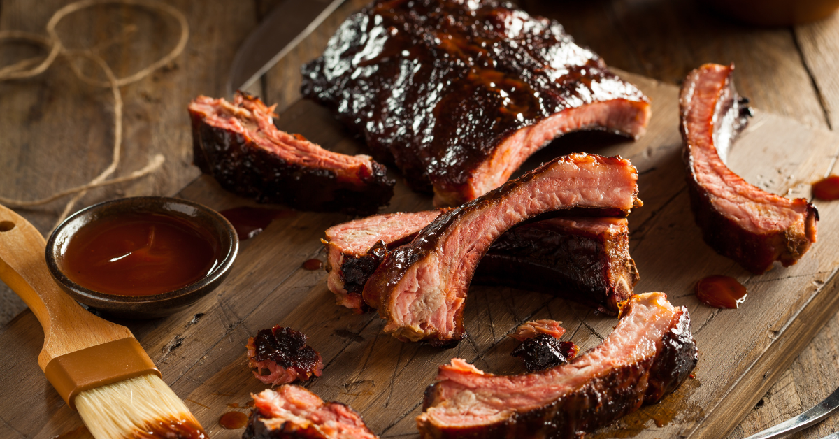 baby back ribs covered in BBQ sauce with a small bowl of BBQ sauce and a basting brush on a wood cutting board