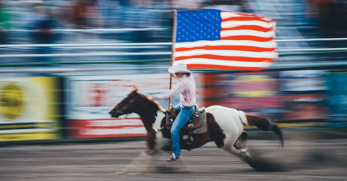 woman riding a paint horse running around a rodeo arena carrying the american flag. woman is wearing blue jeans and a western shirt. horse is brown and white. 