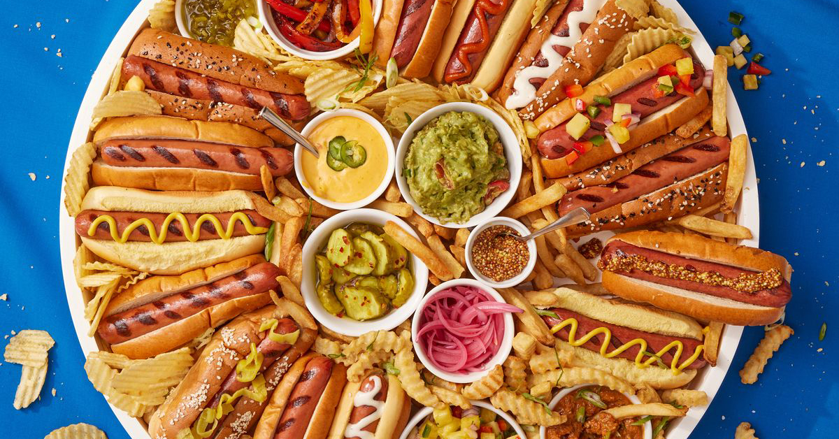 large platter with multiple hot dogs covered in mustard, relish, and peppers with crinkle cut french fries and potato chips around the edges. There are 5 small dishes in the center with onions, pickles, nacho cheese, guacamole and seasonings. 