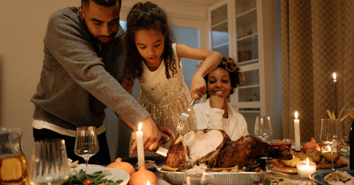 family at thanksgiving dinner table. a woman, man and child. the man and child are carving the thanksgiving turkey. There are wine glasses and candles on the table.