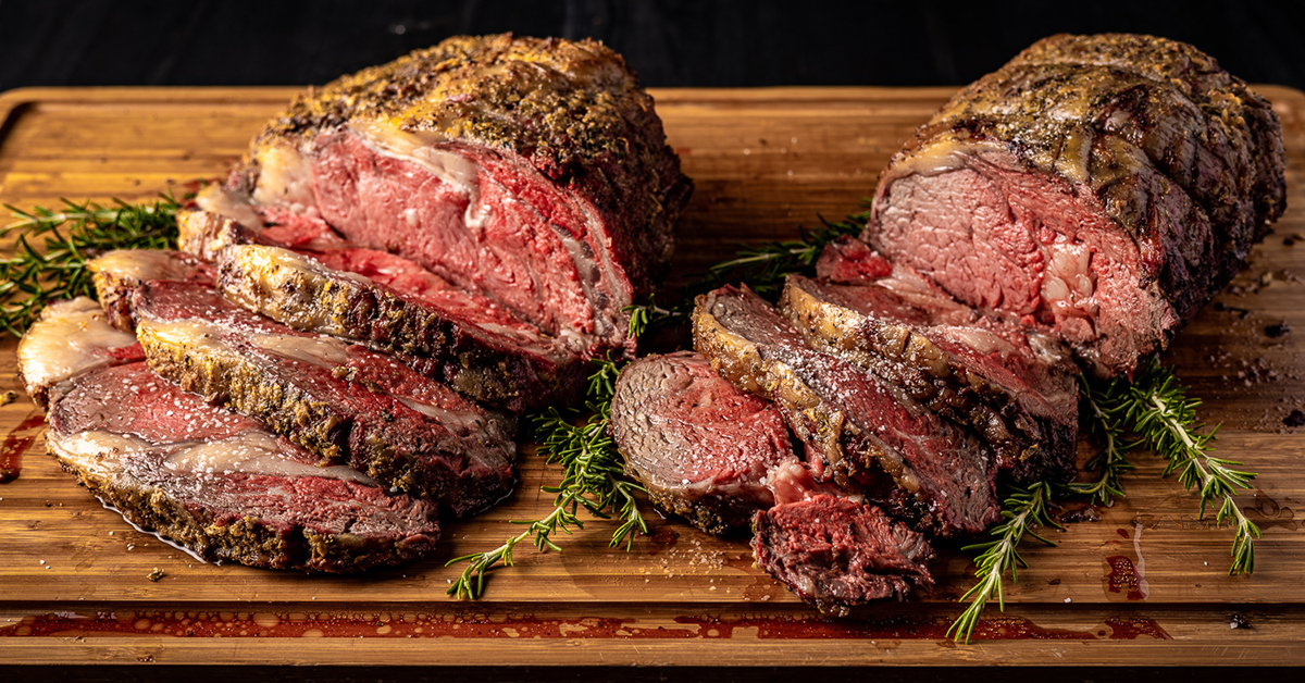 two garlic butter crusted prime ribs on a wooden cutting board, both are sliced half way with three slices on each prime rib. there are rosemary sprigs surrounding the prime ribs.