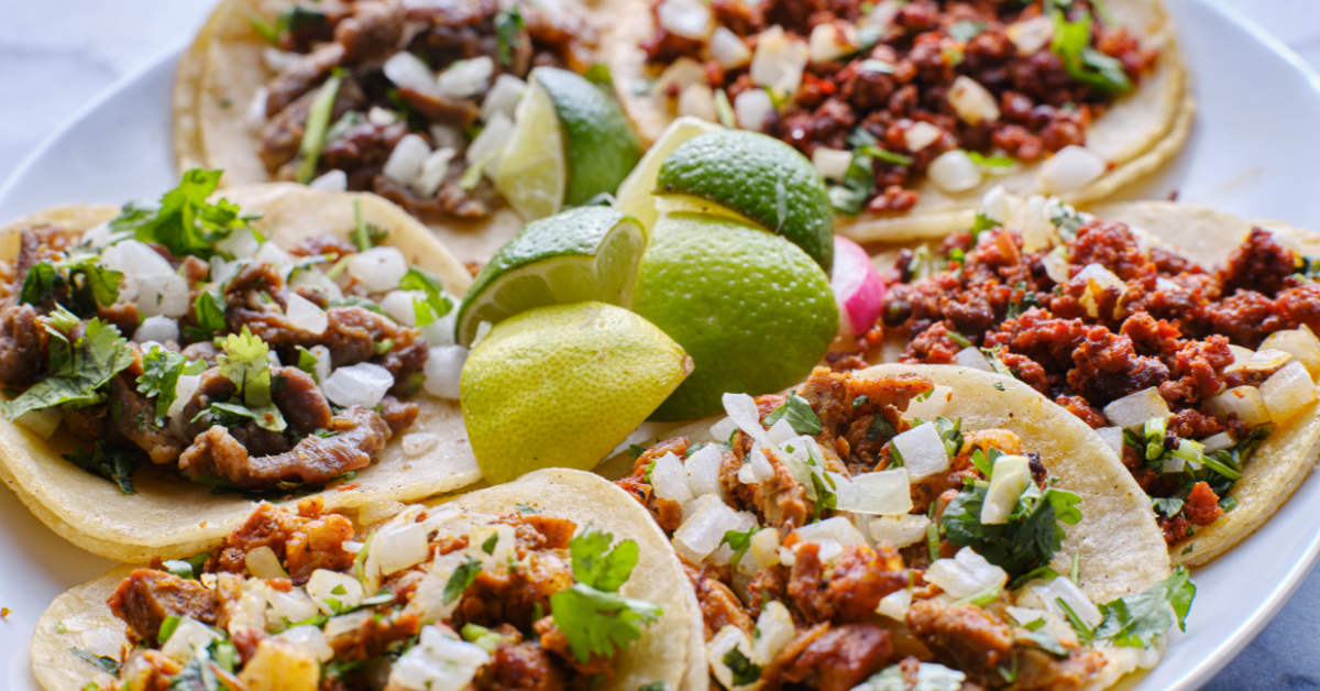 6 tacos on a plate topped with onions and cilantro with 5 sliced limes in the middle