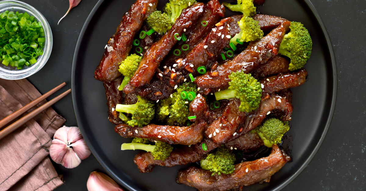 cooked beef and broccoli with sesame seeds on top, on a black plate sitting on a black table with chopsticks and a green garnish on the side