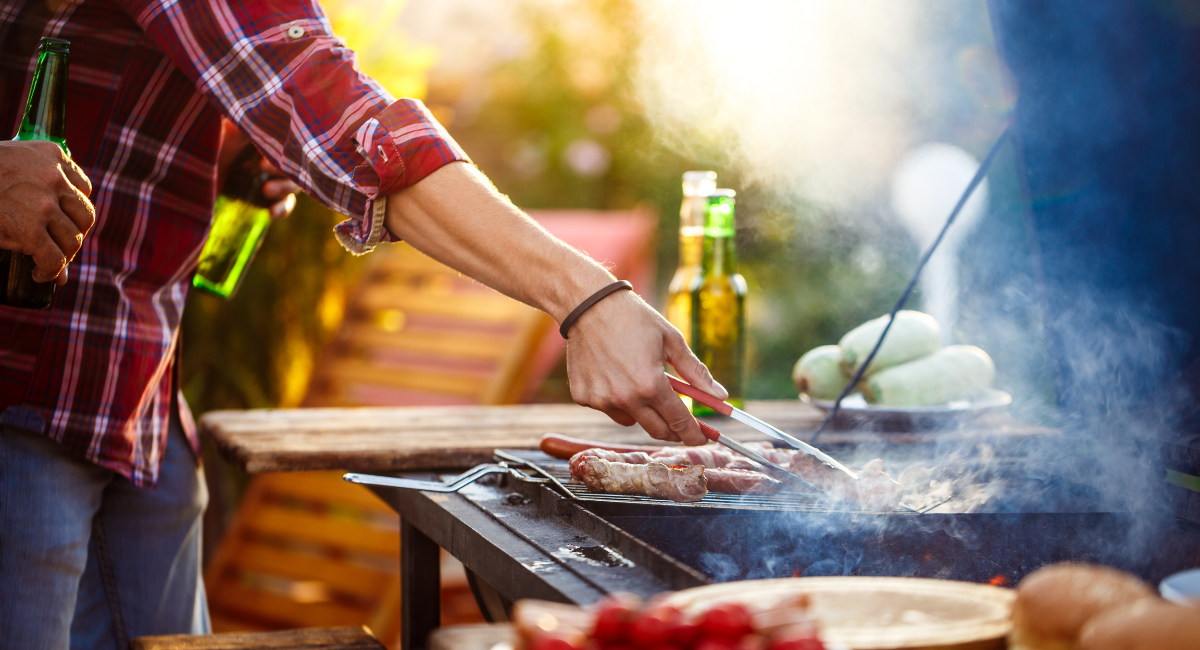 man in flannel flipping a hotdog on the grill, surrounding the grill are vegetables and two beers