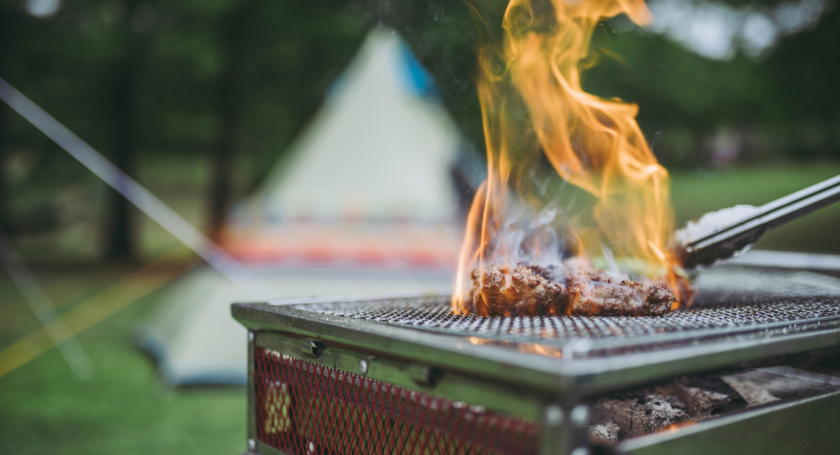 camping essentials for bbq and grilling - photo shows an outdoor grill with a steak and tongs. there is fire surrounding the steak and a tent in the background