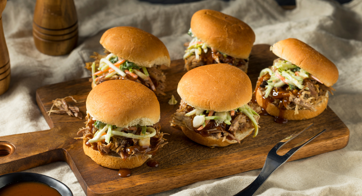 five pulled pork sliders with buns on a wood cutting board with a side of BBQ sauce