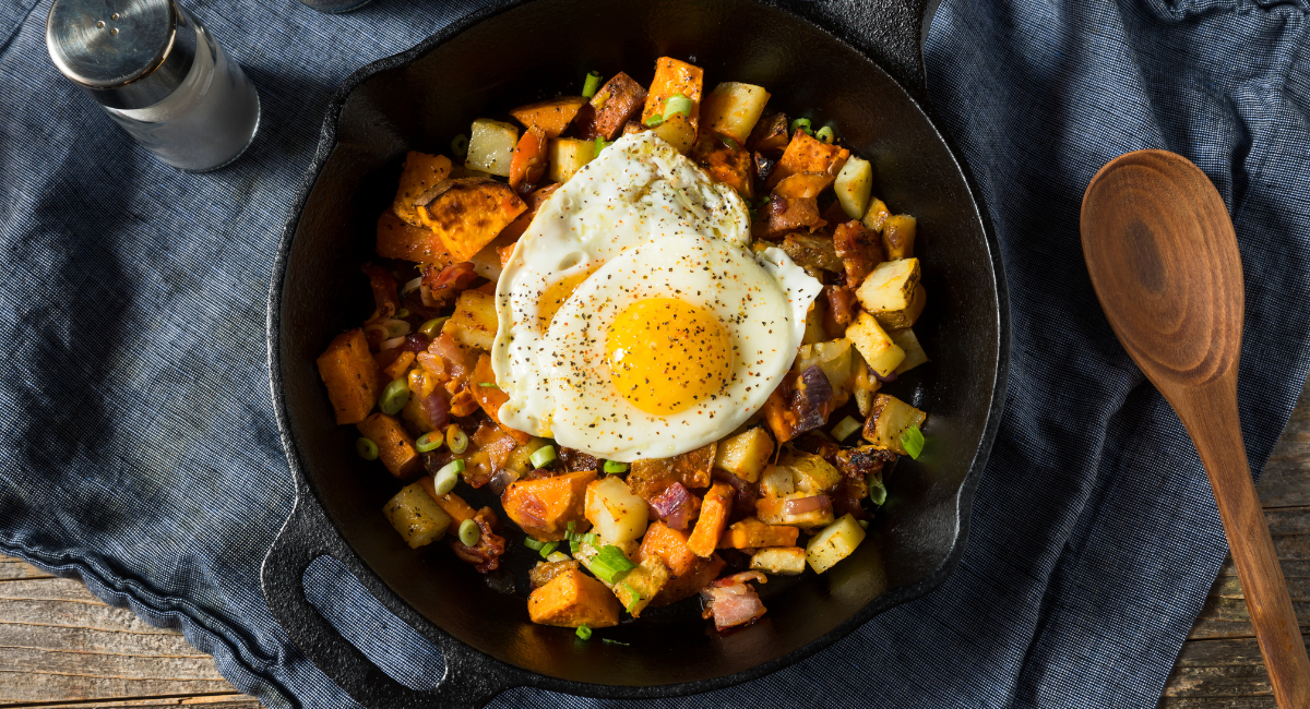 skillet with sausage, potatoes, chives, eggs, salt and pepper. the skillet is placed on a blue cloth and to the right of it is a wooden spoon. to the left of it is a salt shaker filled with salt.
