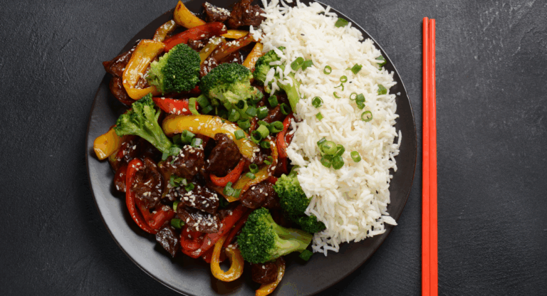 back-to-school dinner idea - a black plate with rice next to sliced beef topped with peppers and broccoli. the plate is on a black table with orange chopsticks next to it