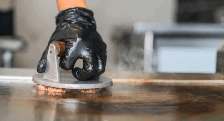 hand with black latex glove on pushing a burger press into a smash burger on a griddle