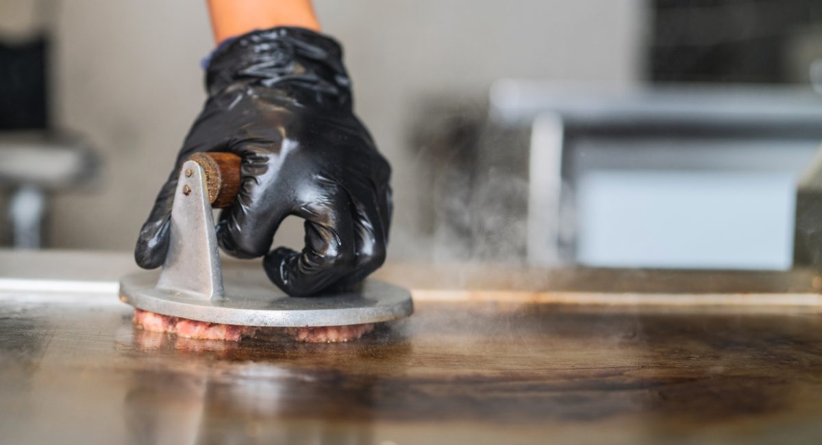 hand with black latex glove on pushing a burger press into a smash burger on a griddle