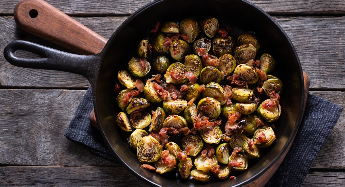 roasted brussels sprouts and small bits of bacon in a cast iron skillet. The skillet is on a wood cutting board, which is on top of a black towel on a wooden table. 