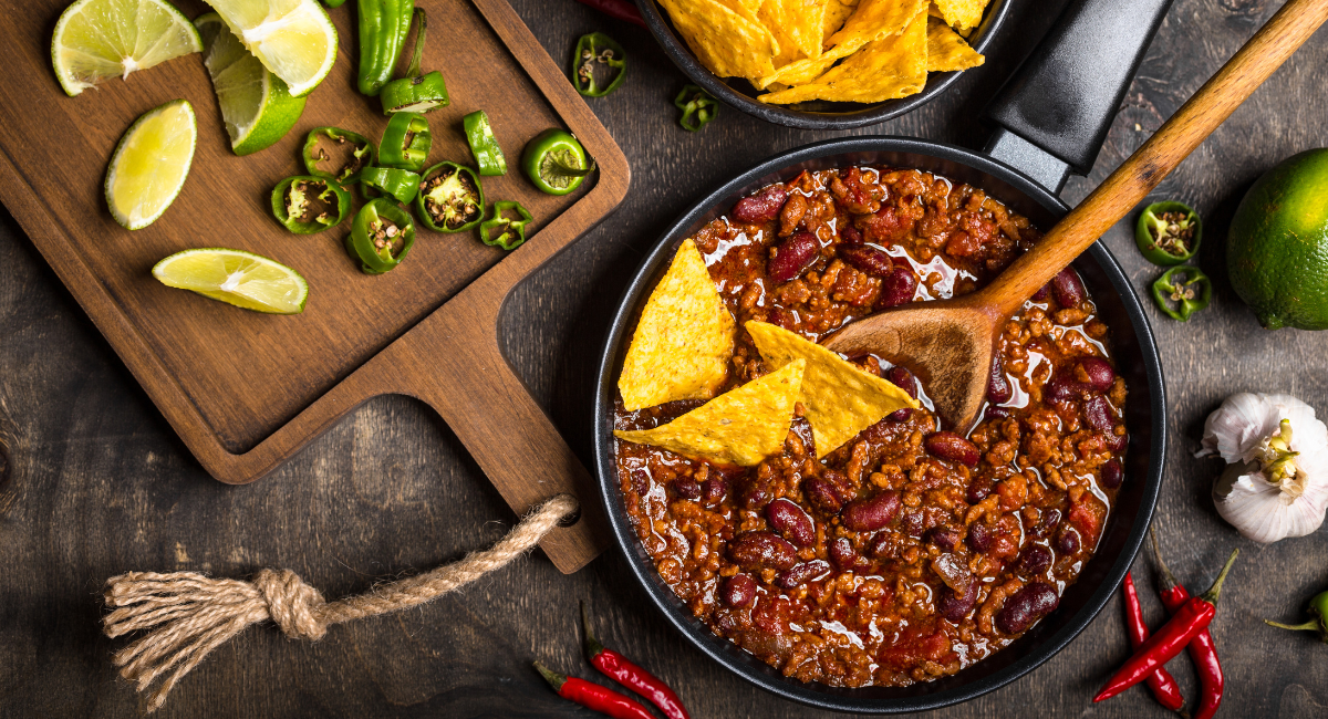 chili in a skillet on a wooden backdrop. next to the chili is a wooden cutting board withs liced limes and jalapenos. there are chili peppers, garlic and limes around the skillet and a bowl of tortilla chips. there are three tortilla chips and a wooden spoon in the chili.