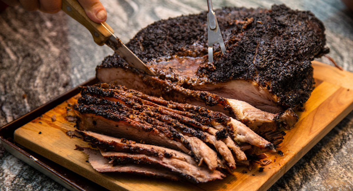 Half-sliced brisket on a wooden cutting board. There are a fork and knife in the brisket actively slicing it