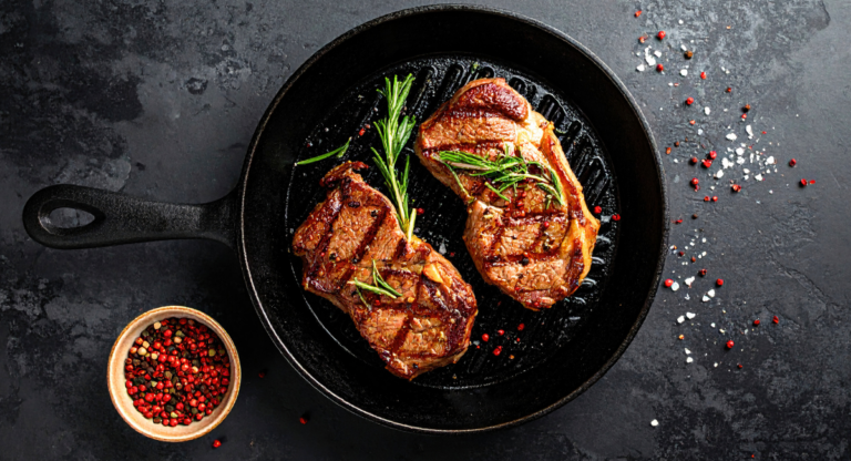 two steaks next to each other on a cast iron skillet. there are sprigs of rosemary on top of the steaks. the skillet is sitting on a black backdrop that has some salt and pepper sprinkled around it
