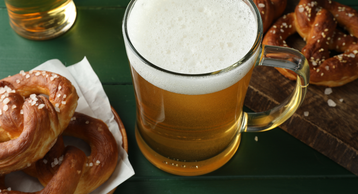 light beer in mug on a green table with a plate of pretzels to the left and two pretzels on a wooden cutting board to the right