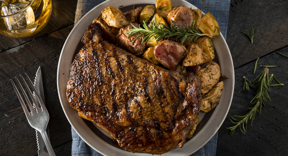 One pot dinner - steak and diced potatoes on a white dish, the white dish is on top of a blue cloth on a wooden table. There is a fork and knife to the left as well as a glass of white wine.