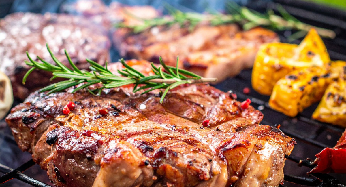 steak on a grill with a sprig of rosemary on top. there are also vegetables in the background