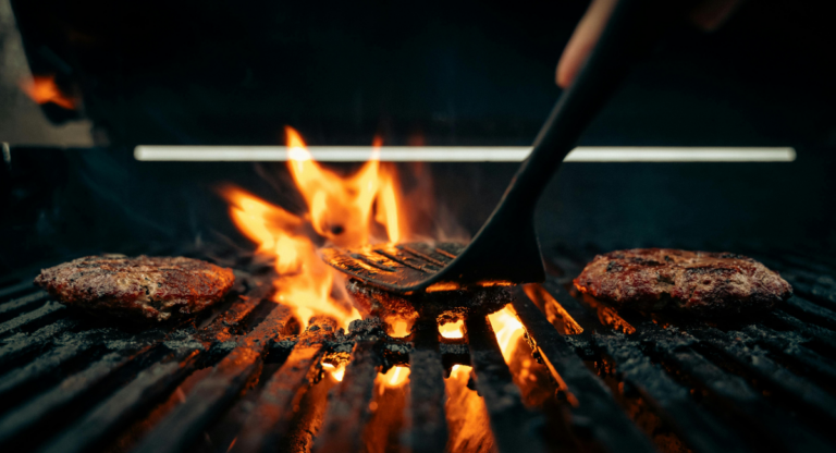 three burgers on a grill with flames coming through the grill grates. there is a spatula smashing down the burger in the middle