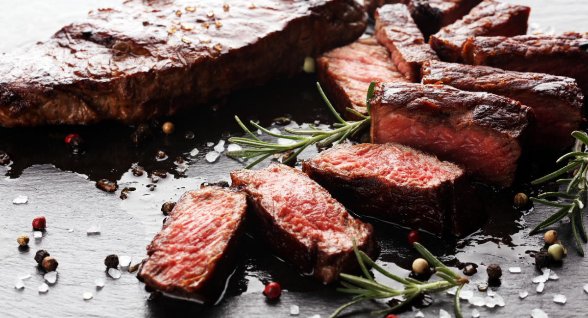 large steak sliced thinly on a black surface. there are peppercorns and rosemary all around the steak