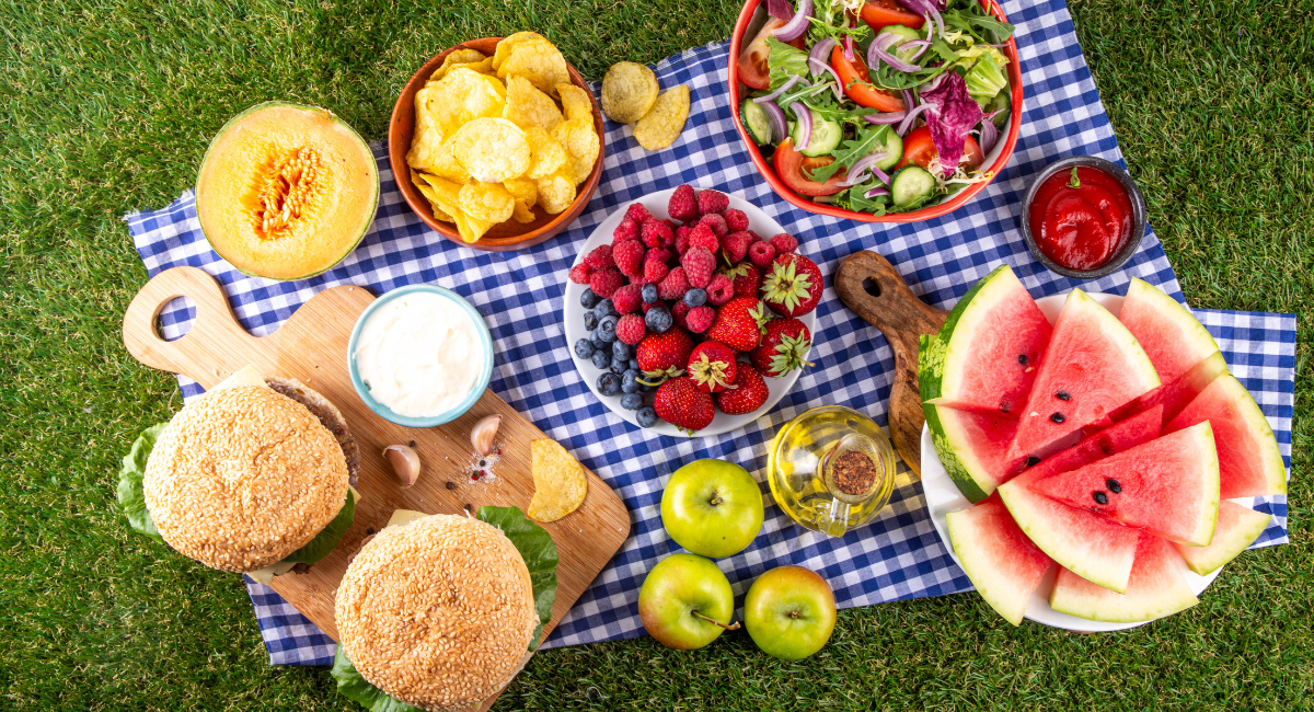 summer produce on a picnic tablecloth - including watermelon, berries, a fresh salad, potato chips, melon, apples and two burgers that are on a cutting board